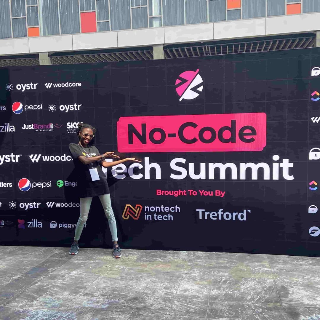 Timonwa is standing in front of the 2022 no-code tech summit banner. She is smiling and pointing to the banner.